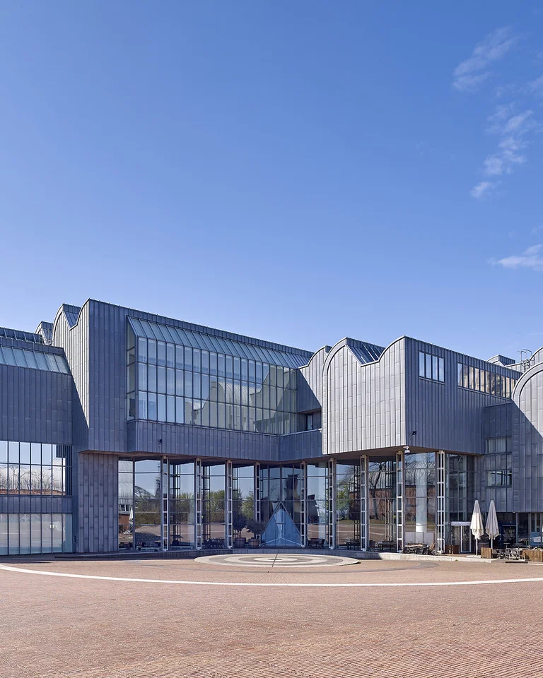 Modern building with large glass panels and a geometric design under a blue sky. Trees and people visible in foreground.