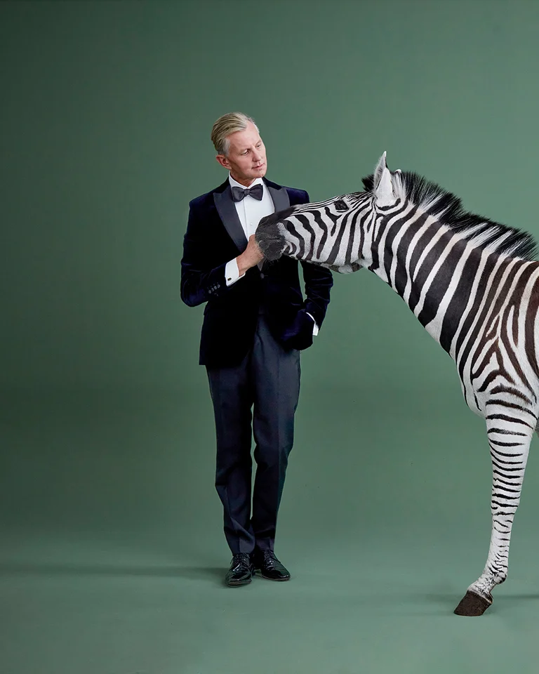 Ein Mann im Smoking steht neben einem Zebra vor einem grünen Hintergrund. Beide scheinen miteinander zu interagieren.