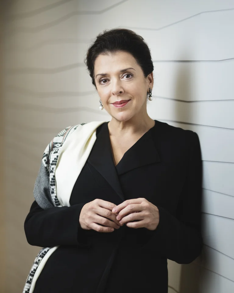 Eine Person steht vor einer Wand, gekleidet in schwarzer eleganter Kleidung und mit einem Schal über der Schulter.