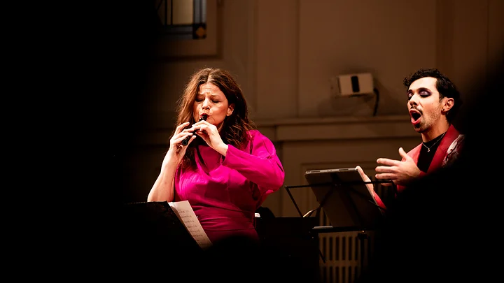 Ein Mann und eine Frau (Ensemble 1700 ) führen klassische Musik in einem Konzertsaal mit dekorierten Fenstern auf. Beide tragen elegante Kleidung.