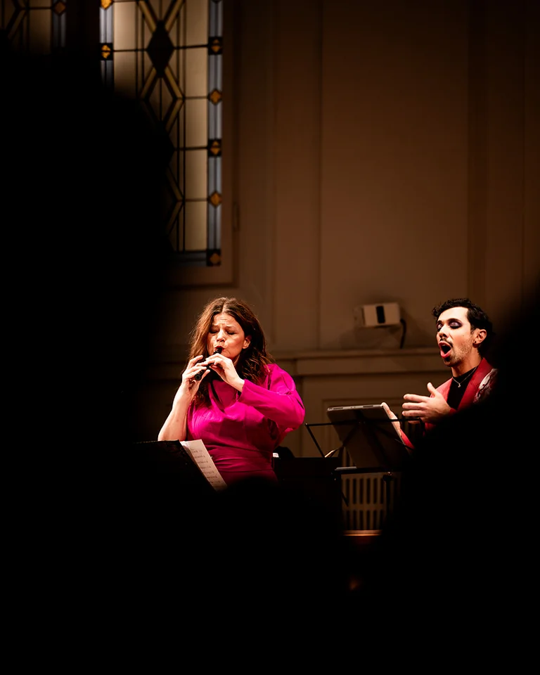 Ein Mann und eine Frau (Ensemble 1700 ) führen klassische Musik in einem Konzertsaal mit dekorierten Fenstern auf. Beide tragen elegante Kleidung.