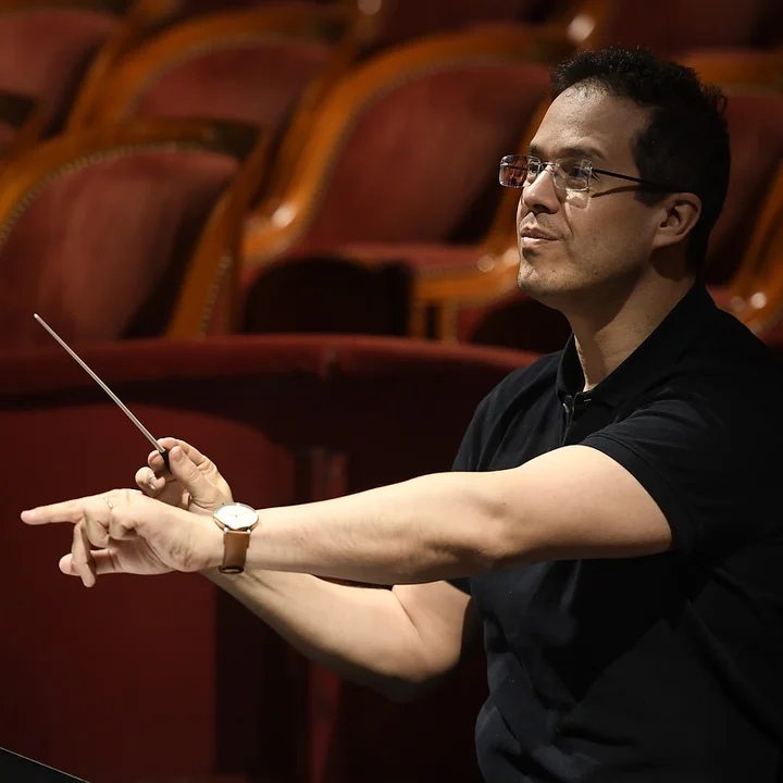 Leonardo García-Alarcón, wearing a dark T-shirt, conducts an orchestra in a hall with rows of red seats.