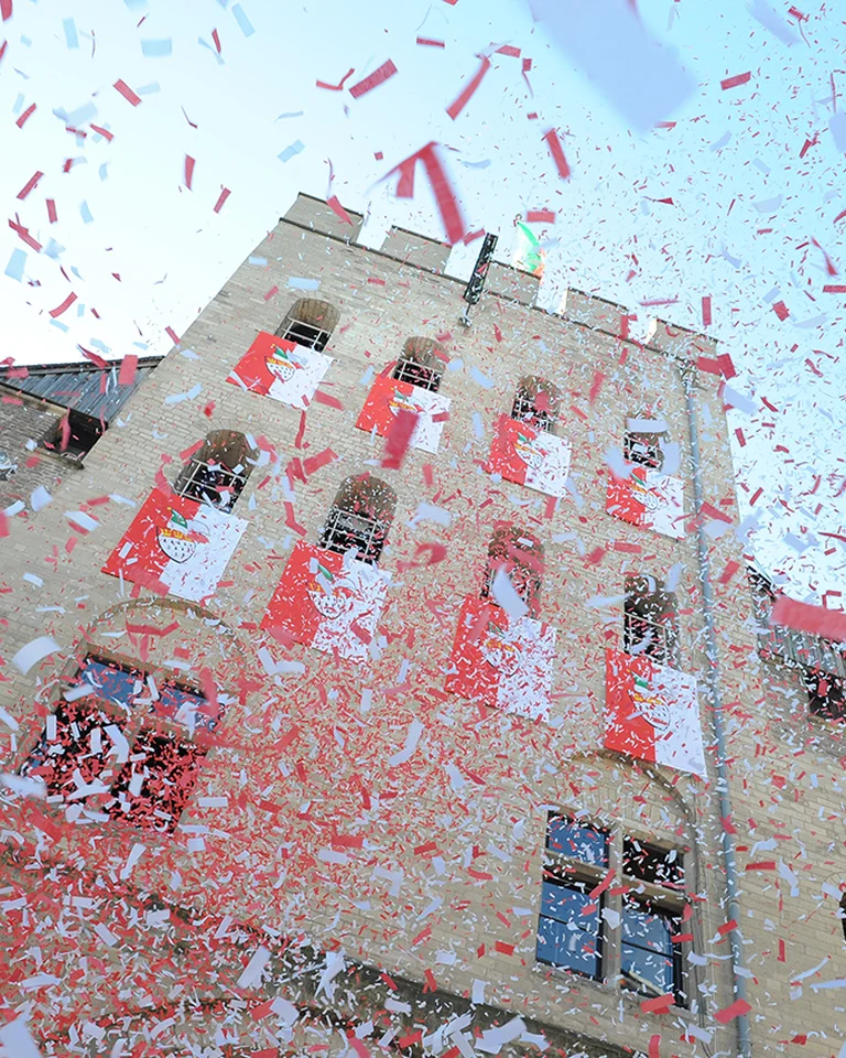 Ein historisches Gebäude mit roten und weißen Bannern, umgeben von herabfallendem Konfetti unter einem klaren, blauen Himmel.