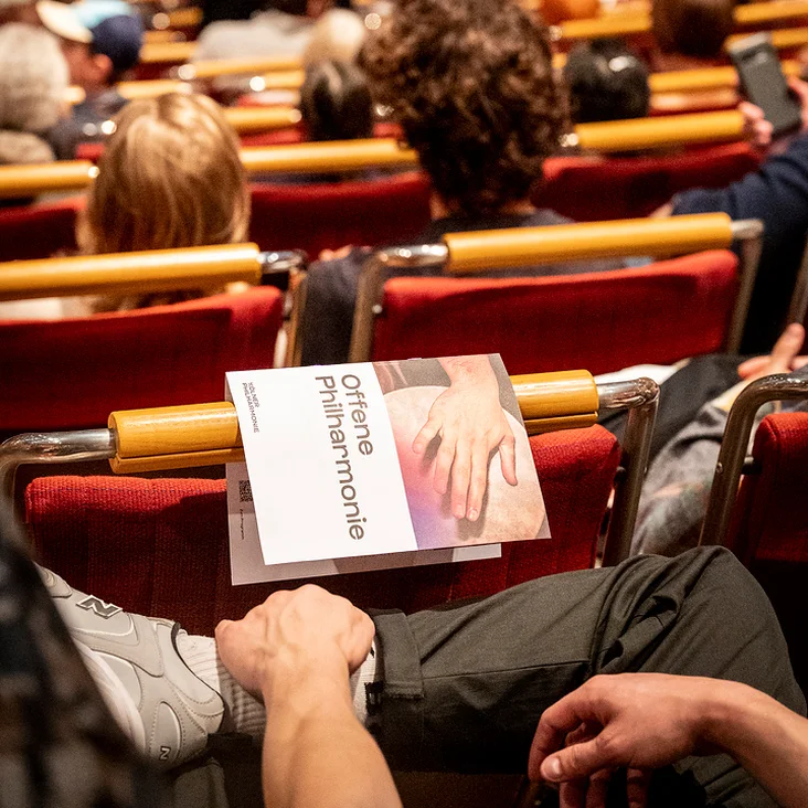Ein Konzertsaal voller Menschen mit einem Programmheft »Offene Philharmonie« auf dem Schoß eines Zuschauers im Vordergrund.
