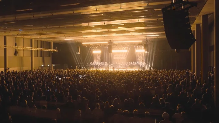 Eine große Menschenmenge bei einem Konzert im stimmungsvoll beleuchteten Saal der Kleinen Olympiahalle. Die Bühne ist voll beleuchtet mit warmem Licht.