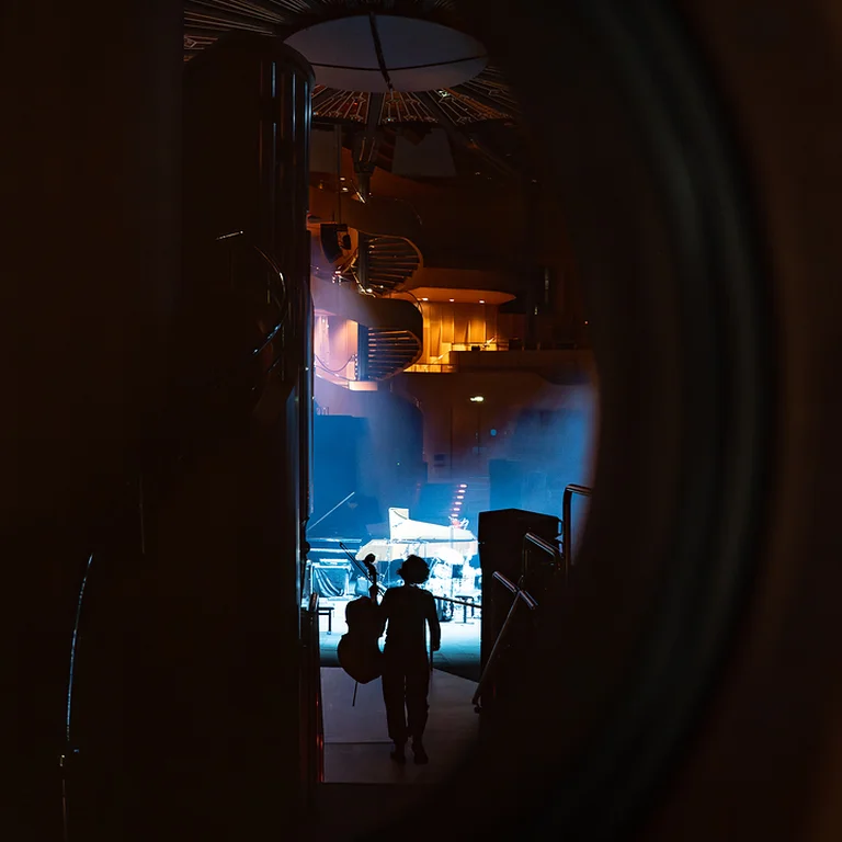 Eine Person (Emily Wittbrodt) steht im Schatten einer Bühne mit blauer und orangefarbener Beleuchtung. Vorbereitungen für eine Aufführung sind im Gange.