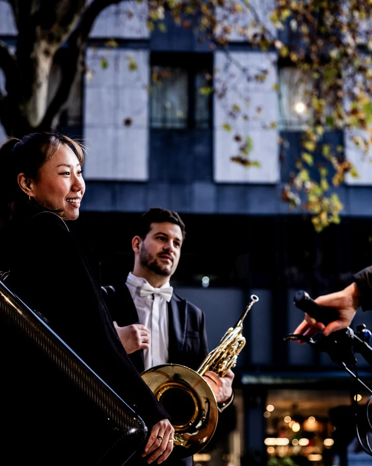 Drei Menschen stehen im Freien. Eine Person hält ein Gehäuse, eine andere hat ein Horn. Es sieht aus wie Abenddämmerung.