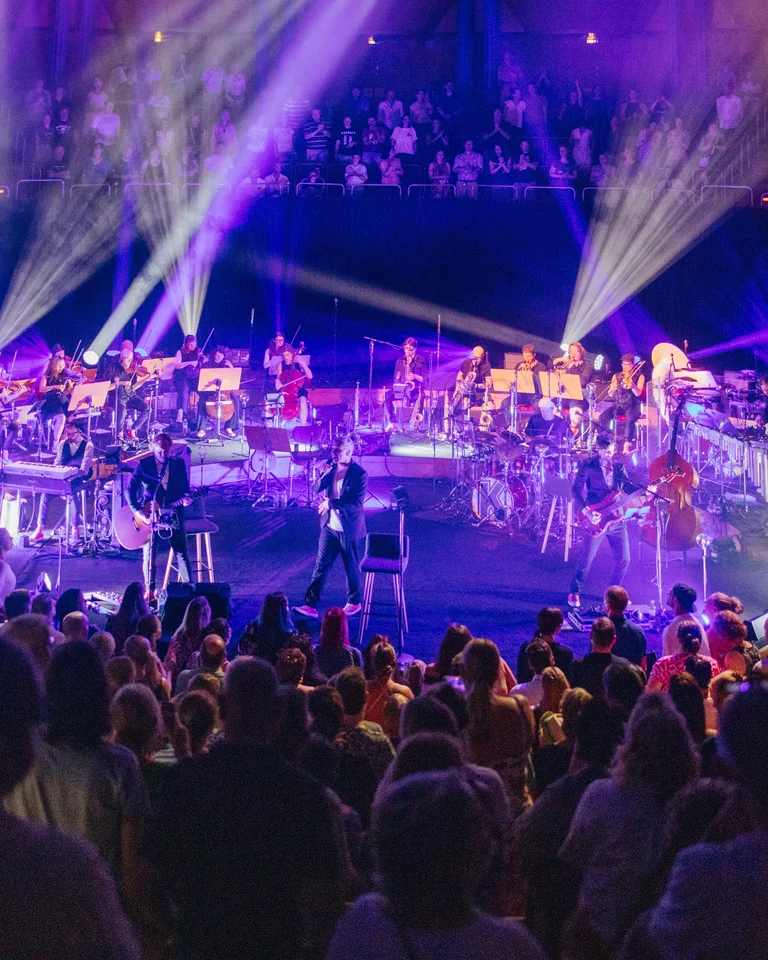 A vibrant stage scene with musicians (Kasalla), bright lights, and an enthusiastic audience in a concert hall. Deep, immersive atmosphere.