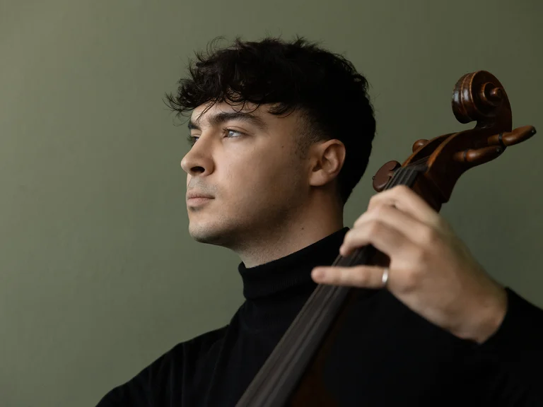 Ein Cellist (Petar Pejčić) in schwarzem Rollkragenpullover spielt konzentriert sein Instrument. Der Hintergrund ist schlicht, was den Fokus auf den Musiker legt.
