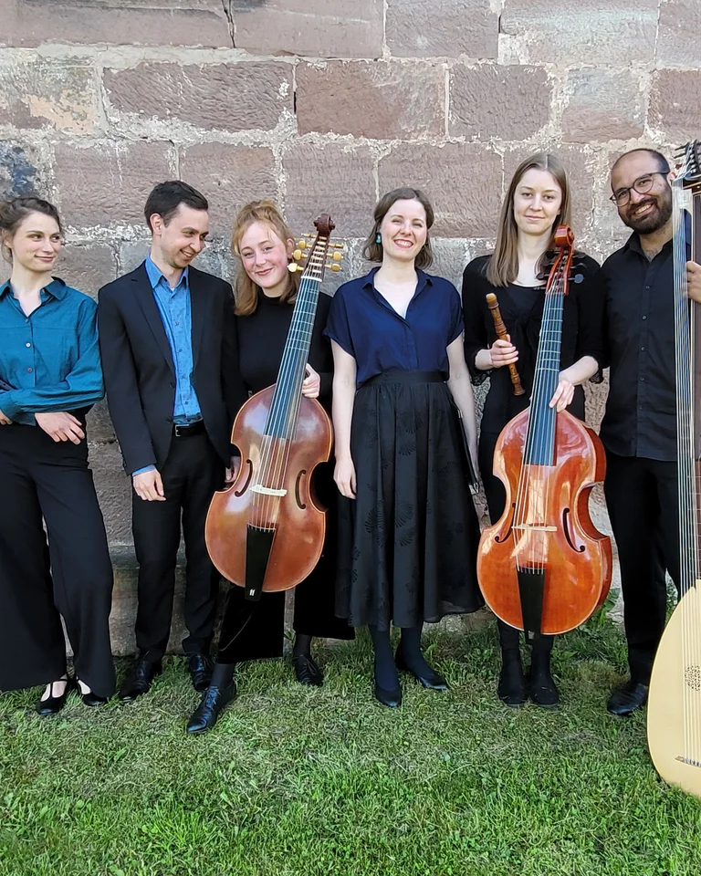 A group of six musicians poses in front of a stone wall. Three hold string instruments; a lute is visible.