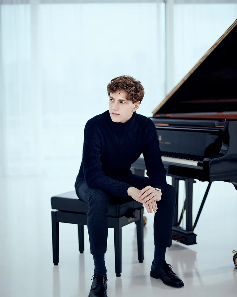 Ein Mann (Jan Lisiecki) sitzt auf einem Hocker neben einem Flügel, in einem minimalistischen, hellen Raum. Er trägt dunkle Kleidung.