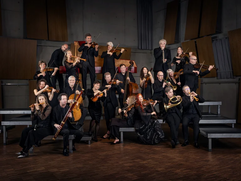 Eine Gruppe von Musikern mit klassischen Instrumenten (Freiburger Barockorchester) posiert auf einer gestuften Bühne in einem modernen Aufführungsraum. Sie tragen schwarze Konzertkleidung.
