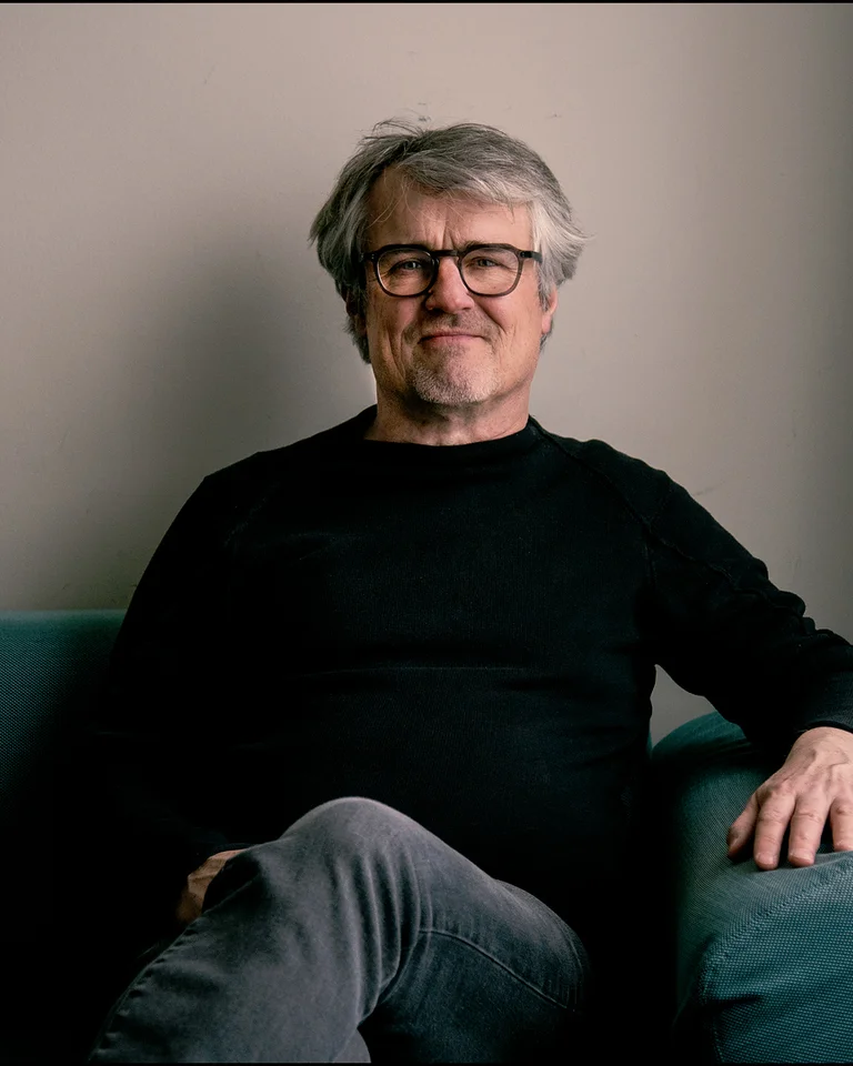 Ein Mann mit Brille (Ingo Metzmacher) sitzt lächelnd auf einem Sofa in einem Raum mit neutralen Farbtönen.