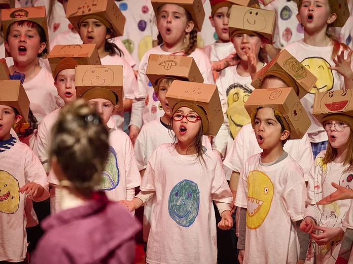 Kinder tragen bemalte T-Shirts und Papphüte, singen auf einer Bühne. Eine Person steht mit dem Rücken zur Kamera.