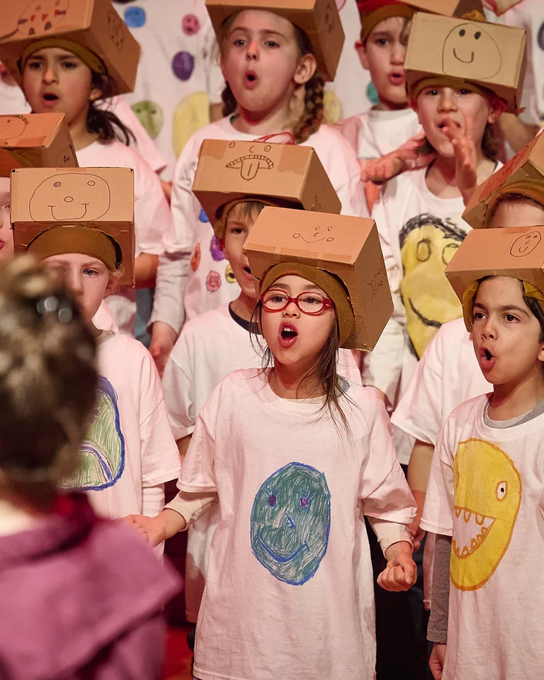 Kinder tragen bemalte T-Shirts und Papphüte, singen auf einer Bühne. Eine Person steht mit dem Rücken zur Kamera.