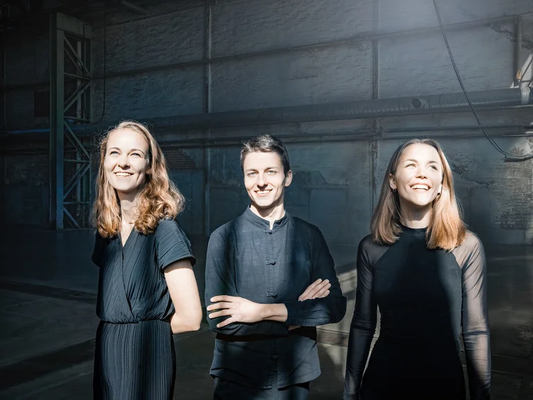 Drei lächelnde Personen (Amelio Trio) in dunkler Kleidung stehen in einem modernen, minimalistischen Raum. Sie wirken freundlich und zuversichtlich.