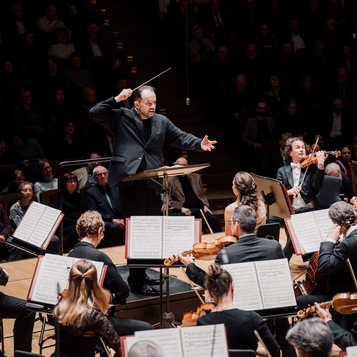 Andris Nelsons mit dem Gewandhausorchester Leipzig: Ein Dirigent, der energisch ein großes Orchester auf einer Bühne dirigiert. Musiker spielen Streich- und Blasinstrumente. Publikum im Hintergrund.