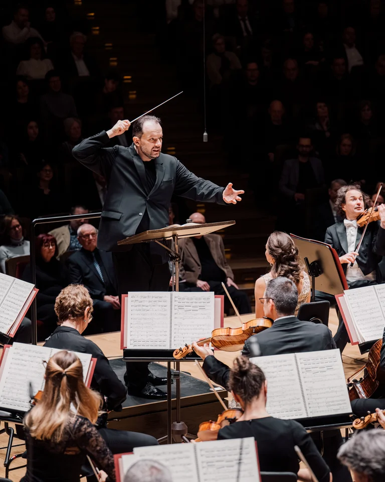 Andris Nelsons mit dem Gewandhausorchester Leipzig: Ein Dirigent, der energisch ein großes Orchester auf einer Bühne dirigiert. Musiker spielen Streich- und Blasinstrumente. Publikum im Hintergrund.
