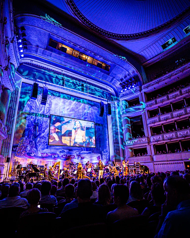 Rock the Opera: A lit theater with an audience, orchestra on stage. The space is bathed in blue light, large screen in the background.