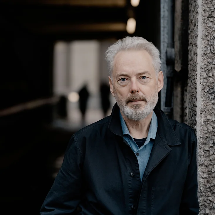 Ein Mann (Mark Padmore) mit grauem Haar und Bart steht in einem dunklen Gang und trägt eine schwarze Jacke, im Hintergrund unscharfe Silhouetten.