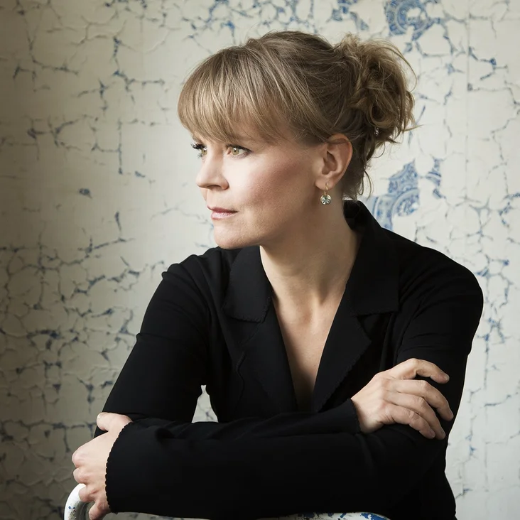 Eine Frau mit blonden Haaren und einem schwarzen Oberteil sitzt und schaut nach rechts vor einer strukturierten Wand.