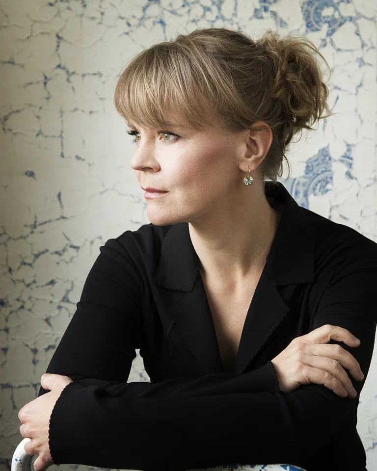 Eine Frau mit blonden Haaren und einem schwarzen Oberteil sitzt und schaut nach rechts vor einer strukturierten Wand.