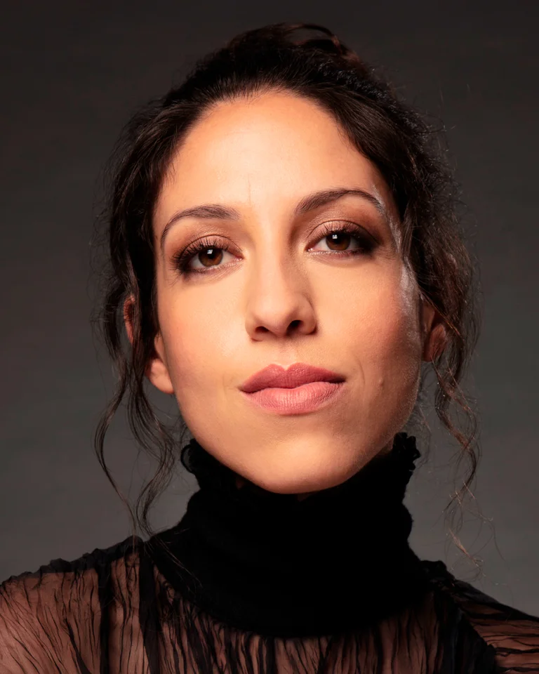 Eine Frau mit dunklem Haar trägt ein schwarzes, halbtransparentes Oberteil vor einem neutralen Hintergrund und schaut nachdenklich.