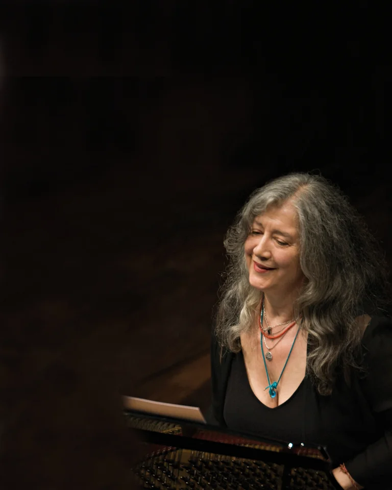 A woman with long gray hair plays the piano with focus. She wears a black top and colorful necklace.