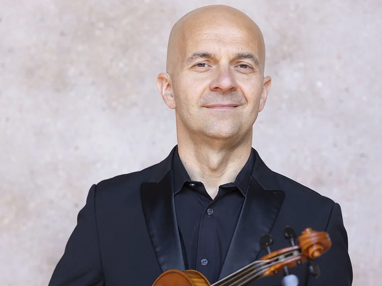 Ein Mann (Alessandro Tampieri) in einem eleganten Anzug hält eine Geige. Er steht vor einem neutralen Hintergrund und lächelt.