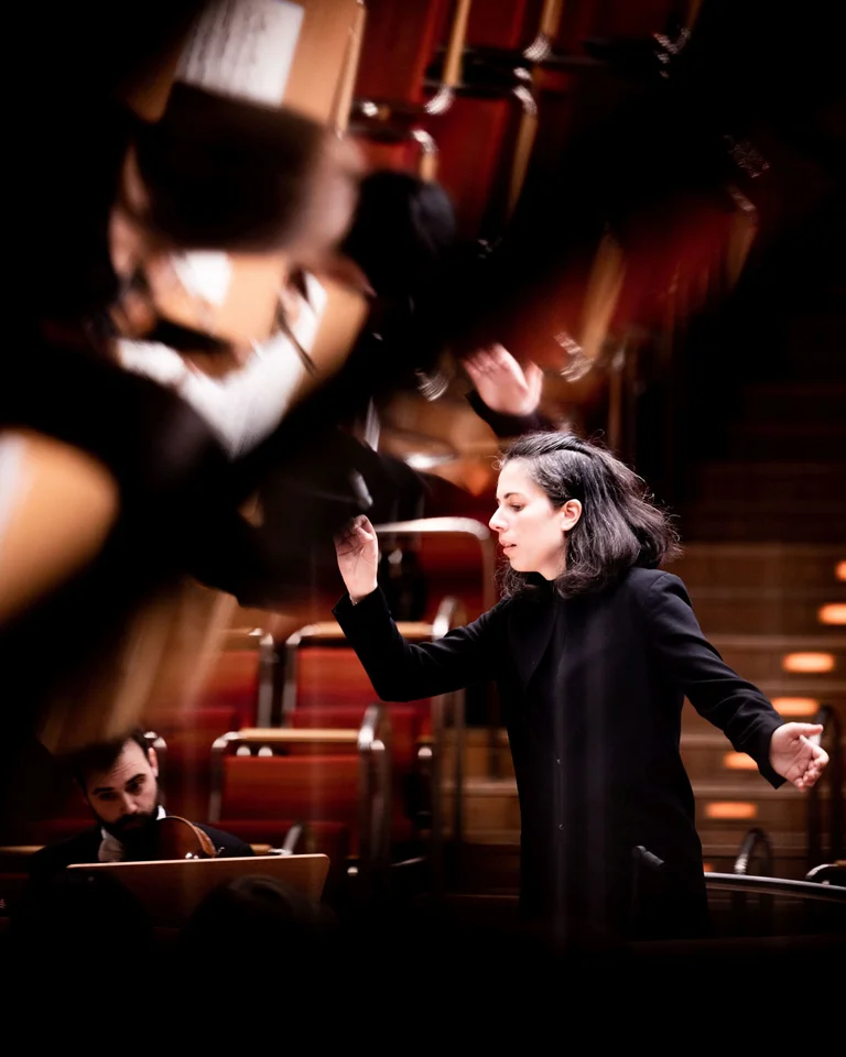Eine Dirigentin leitet intensiv ein Orchester in einem Konzertsaal. Im Hintergrund sind gestaffelte Sitzreihen zu sehen.
