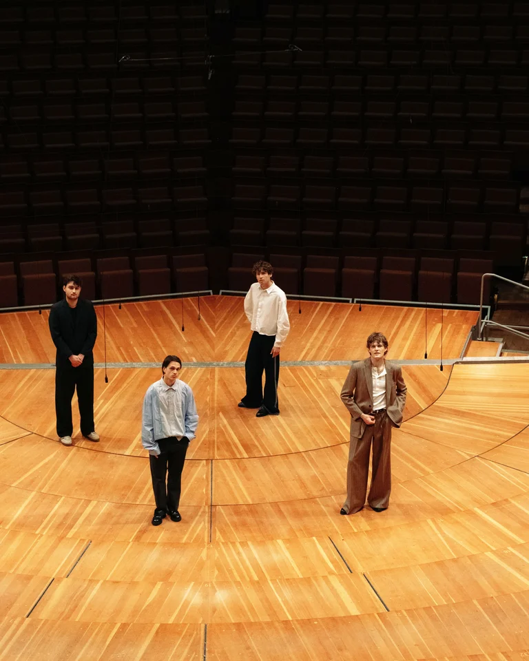 Vier Personen (JEREMIAS) stehen auf einer großen, leeren Bühne in einem Konzertsaal. Die Bühne ist aus hellem Holz gefertigt.