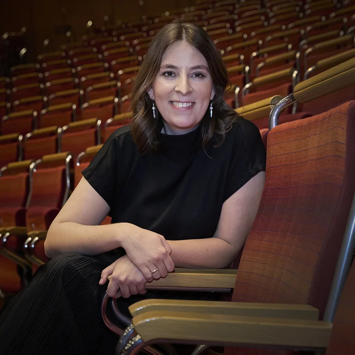 Charlotte Oelschlegel sitzt in einer Stuhlreihe im leeren Konzertsaal der Kölner Philharmonie. Sie trägt schwarze Kleidung und lächelt in die Kamera.