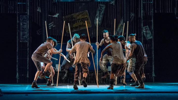 Eine Gruppe von acht Tänzern auf der Bühne, die dynamisch mit Stöcken in synchronisierter Choreografie agieren, vor einem urbanen Hintergrund.