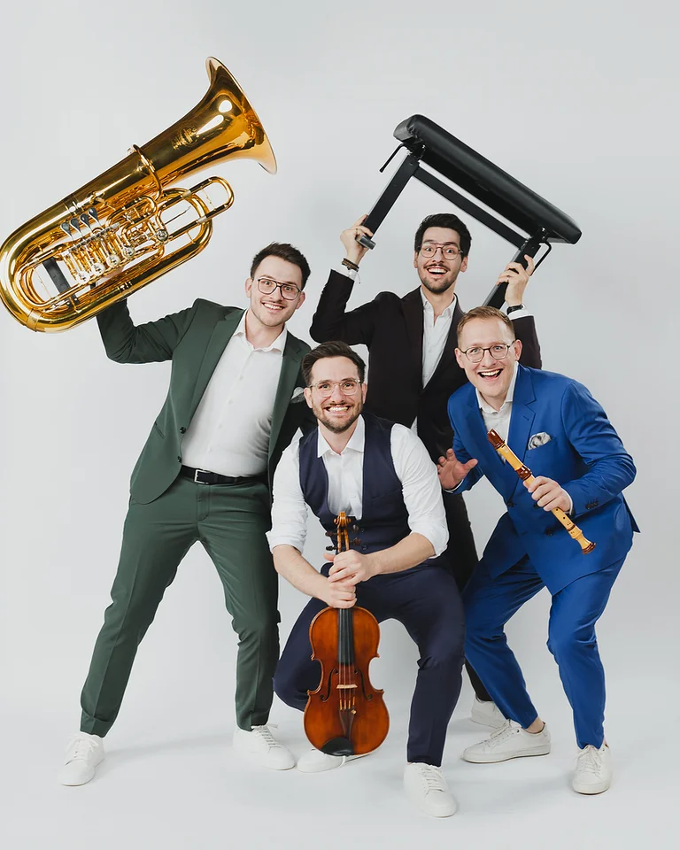 Vier Männer (Hanke Brothers) posieren fröhlich mit Musikinstrumenten: einer spielt Cello, die anderen zeigen Tuba, Trommel und Flöte.