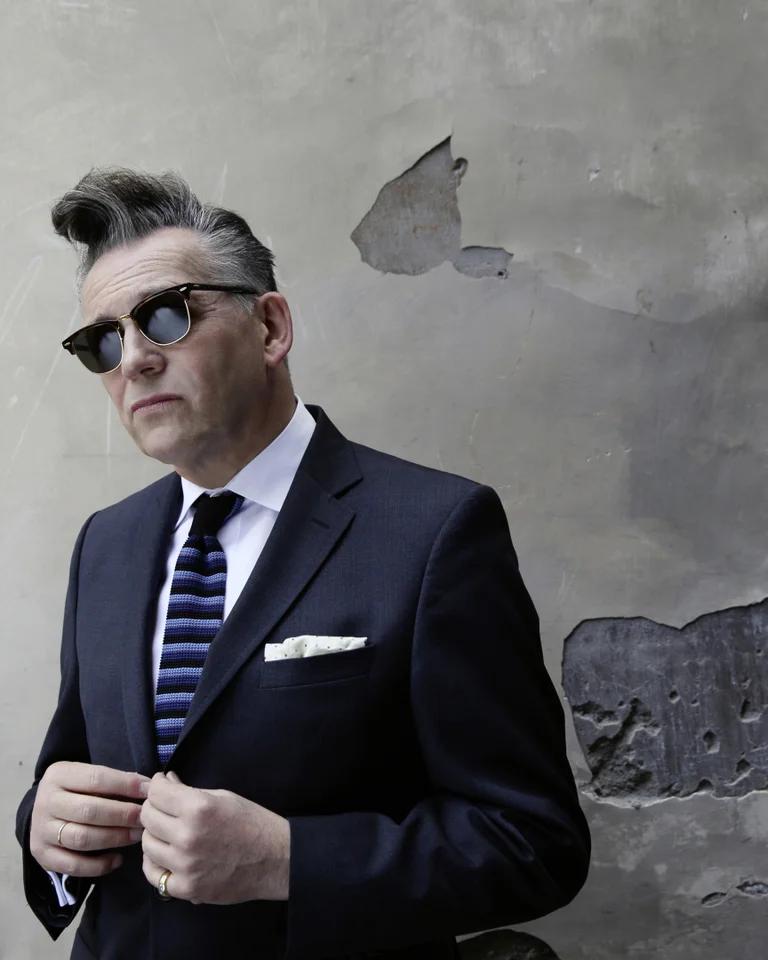 A man in a suit with sunglasses stands in front of a worn wall, looking to the side. Stylish and classic.