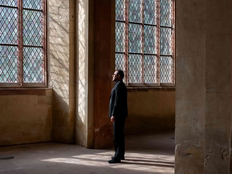 Ein Mann (Andreas Scholl) steht in einer Kirche mit großen, gotischen Fenstern. Sonnenlicht fällt auf den Boden und erzeugt ein Gefühl der Ruhe.