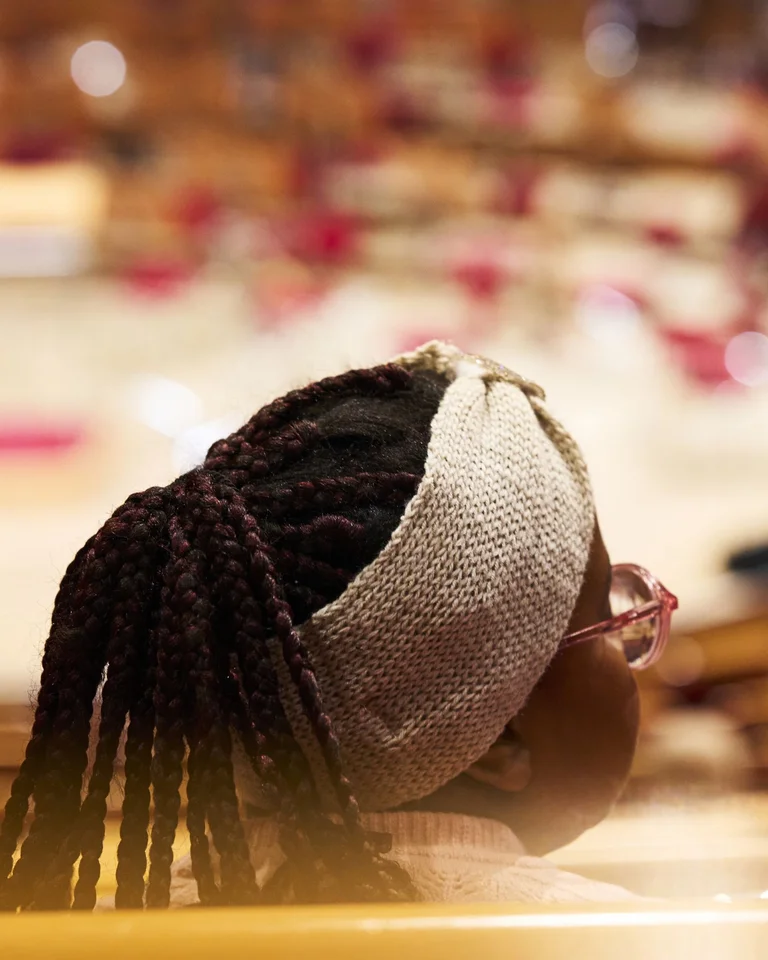 Close-up of a person with braided hair and a headband watching in a concert hall, with blurred chairs and orchestra in the background.