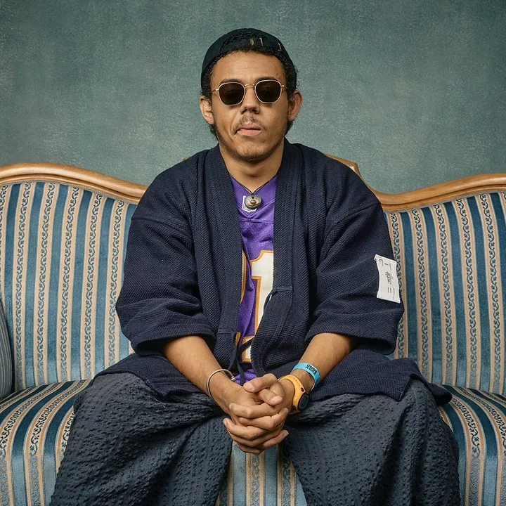 Emilio Modeste is sitting on a striped sofa, wearing sunglasses, a baseball cap, and comfortable, sporty clothing.