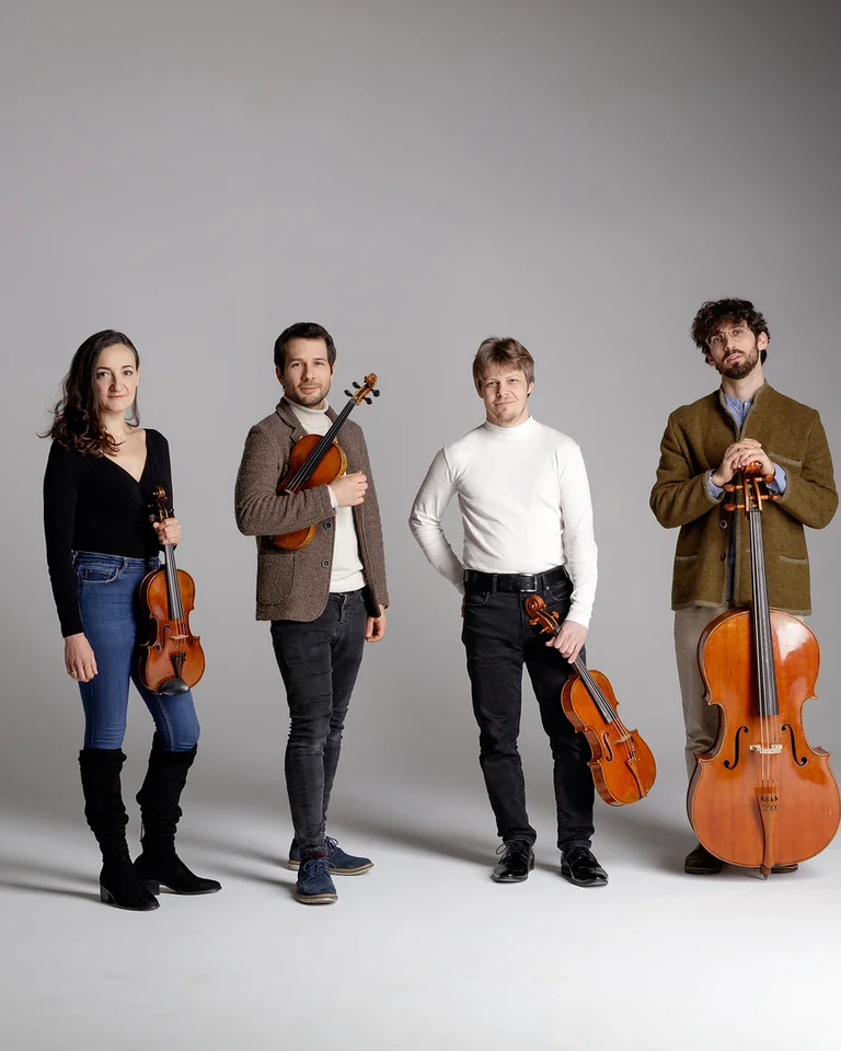 Vier Musiker stehen mit ihren Streichinstrumenten auf einem neutralen Hintergrund. Sie tragen lässige, moderne Kleidung und blicken in die Kamera.