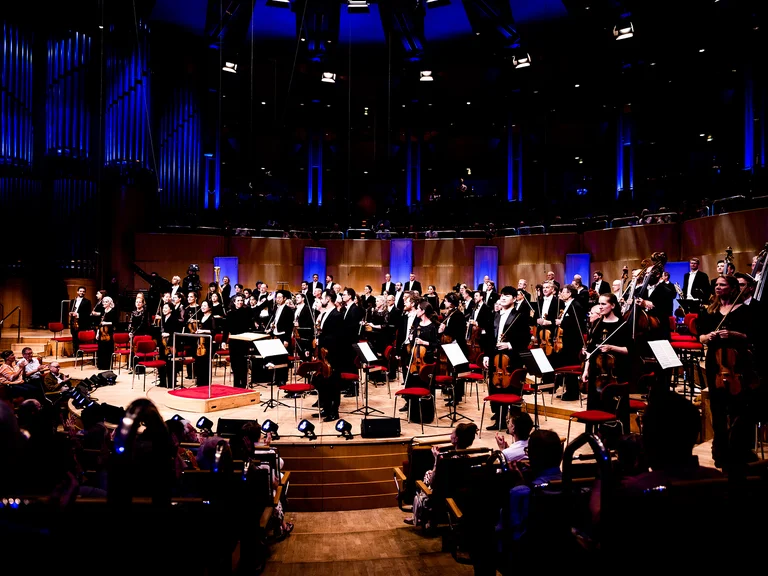 Ein großes Orchester (WDR Sinfonieorchester) spielt in einem beleuchteten Konzertsaal mit Publikum. Die Musiker stehen, um Applaus zu empfangen.