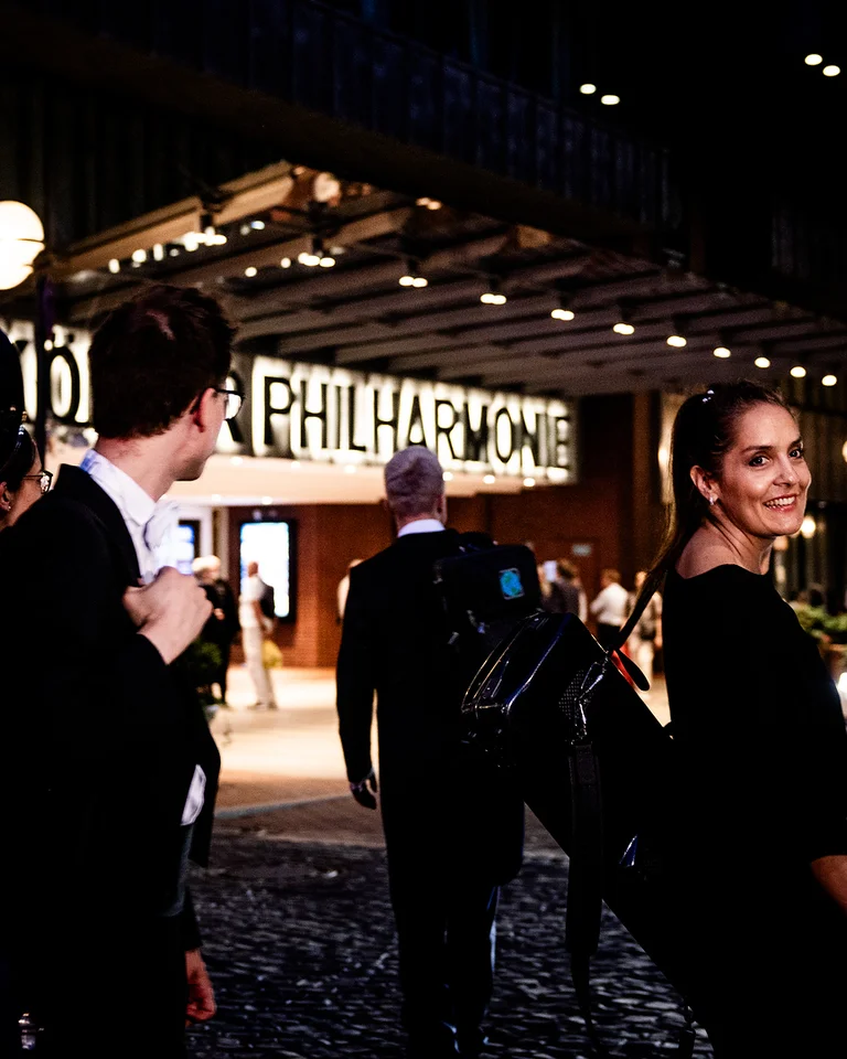 MusikerInnen (WDR Sinfonieorchester) in Abendkleidung betreten lächelnd die Kölner Philharmonie mit heller Beleuchtung..