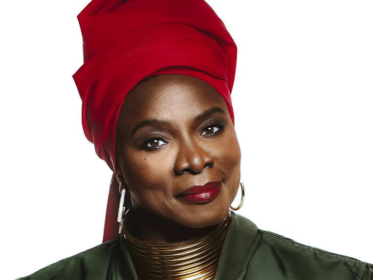 Frau (Angélique Kidjo) mit rotem Kopftuch und goldenen Halsreifen, trägt eine grüne Jacke, posiert selbstbewusst vor einem schlichten weißen Hintergrund.
