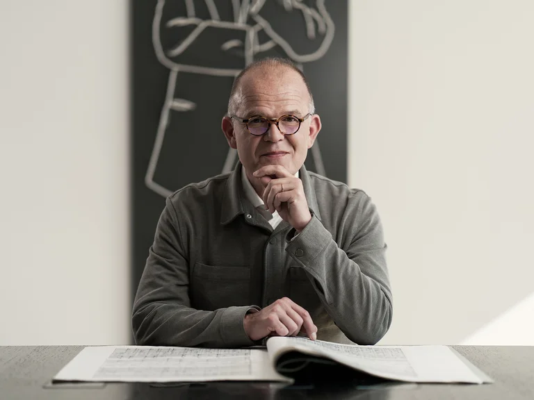 Ein Mann (François-Xavier Roth) sitzt an einem Tisch mit geöffnetem Partitur, im Hintergrund eine Tafelzeichnung einer Person.