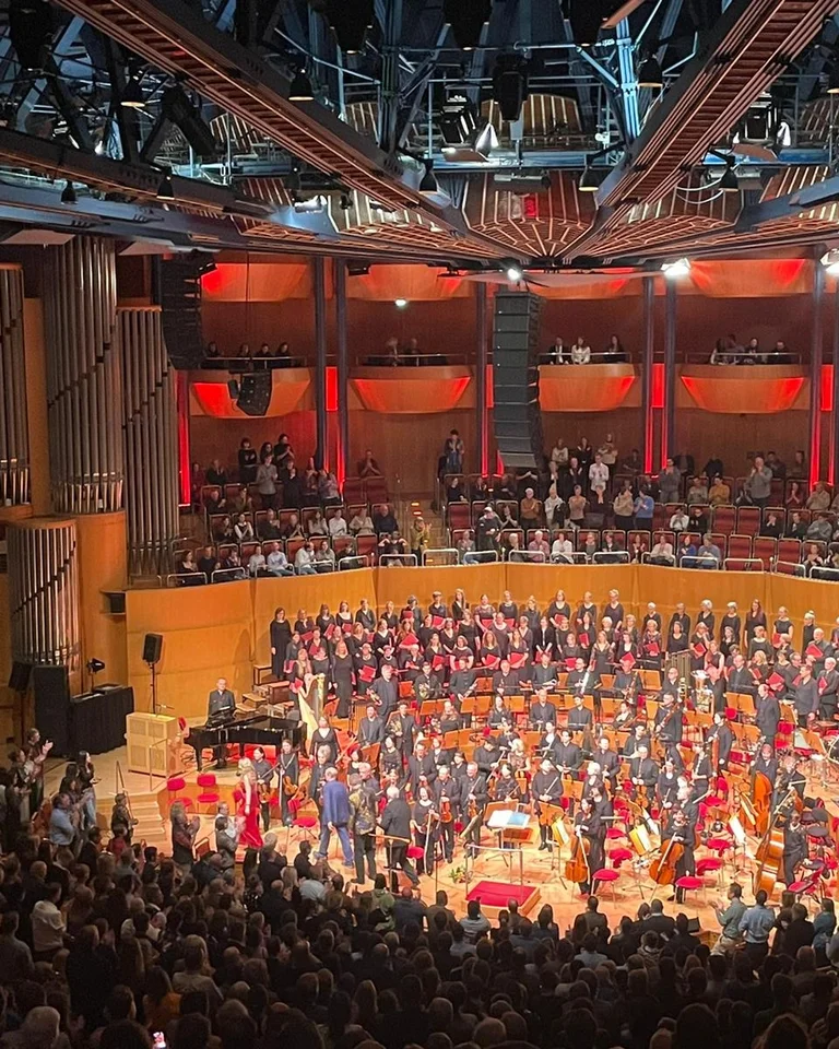 Ein großes Orchester und ein Chor (KölnChor) vor einem vollen Publikum in einem modernen Konzertsaal (Kölner Philharmonie) mit beeindruckender Architektur und stimmungsvoller Beleuchtung.