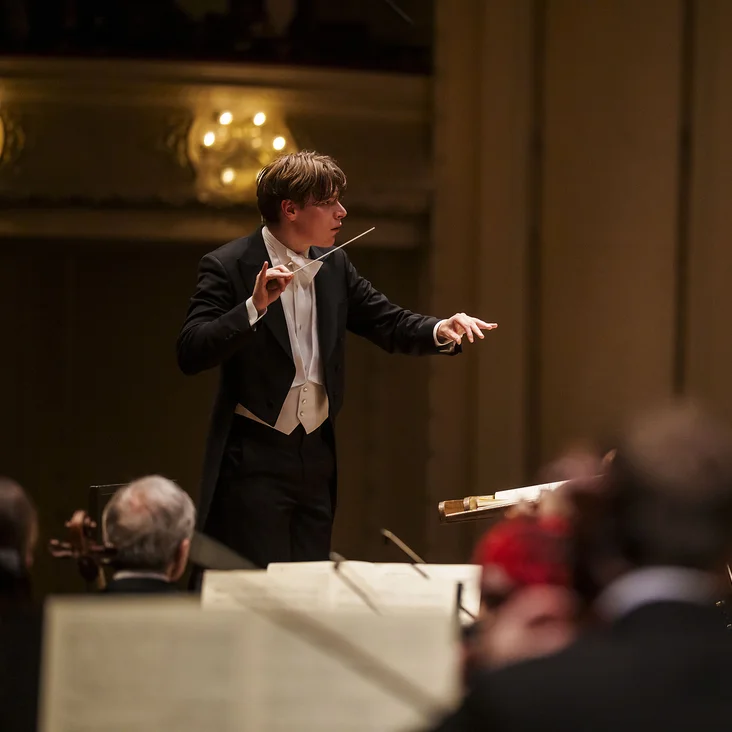 Ein Dirigent (Klaus Mäkelä) leitet ein Orchester (Chicago Symphony Orchestra) in einem Konzertsaal. Musiker spielen Geigen, Celli und andere Instrumente. Die Bühne ist elegant beleuchtet.
