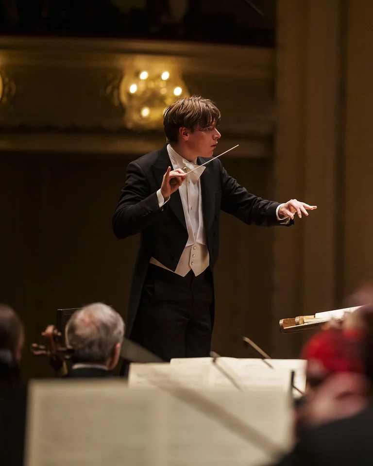 Ein Dirigent (Klaus Mäkelä) leitet ein Orchester (Chicago Symphony Orchestra) in einem Konzertsaal. Musiker spielen Geigen, Celli und andere Instrumente. Die Bühne ist elegant beleuchtet.