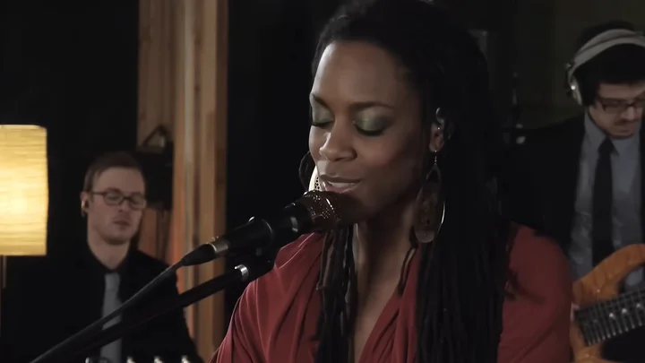 Akua Naru sings into a microphone with her eyes closed. Band members with instruments can be seen in the background.