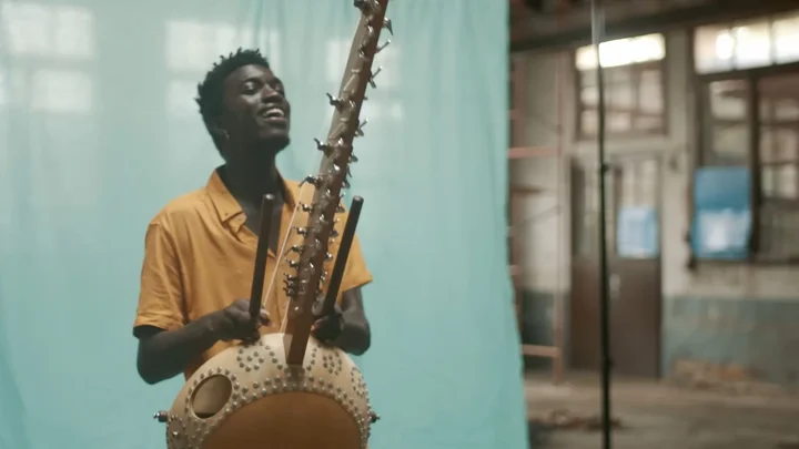 Momi Maiga spielt eine afrikanische Kora vor einem türkisfarbenem Vorhang in einem leerstehenden Industriegebäude.