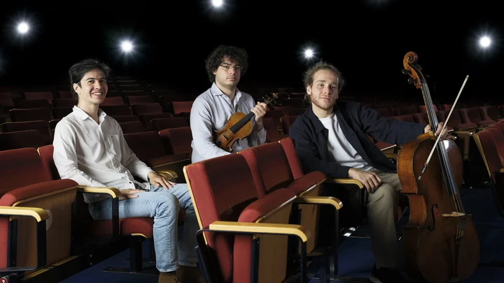 The three members of Trio Concept are sitting in the rows of seats in the empty hall of the Kölner Philharmonie. Two of them are holding their musical instruments: a violin and a cello.