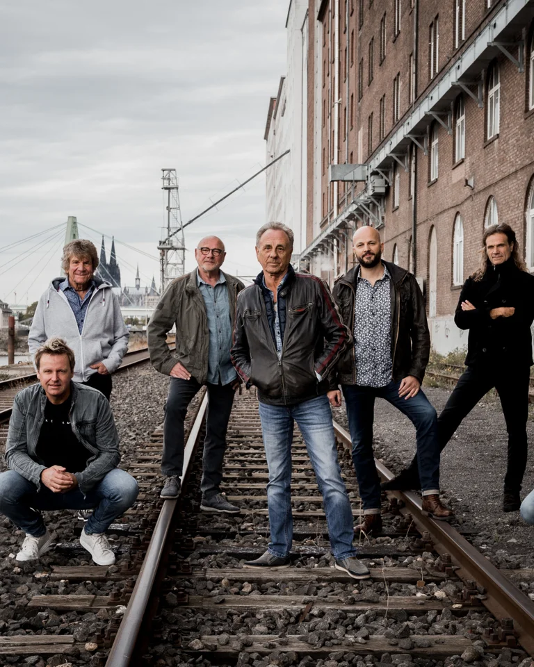 A group of eight people casually posing on old railway tracks beside a brick building. The sky appears cloudy.